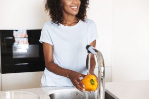 A young, smiling woman stands at her kitchen sink washing a yellow pepper with tapwater.
